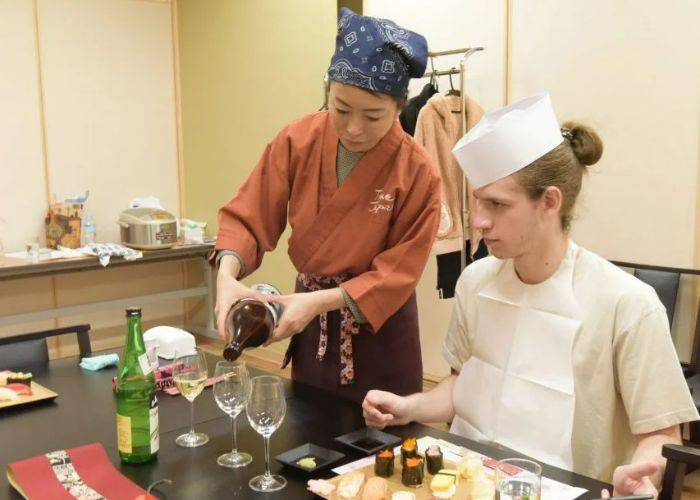 A host pouring a glass of sake for a guest. In front of them, a plate is filled with different Japanese foods.